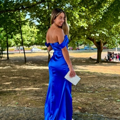 Mulher de vestido azul royal longo no parque, clutch branca, óculos redondos.