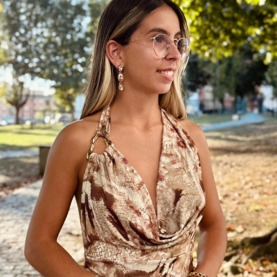 Mulher com vestido estampado castanho e bege, óculos e acessórios dourados com parque ao fundo