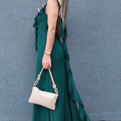 Mulher com vestido verde longo e mala bege contra parede cinza