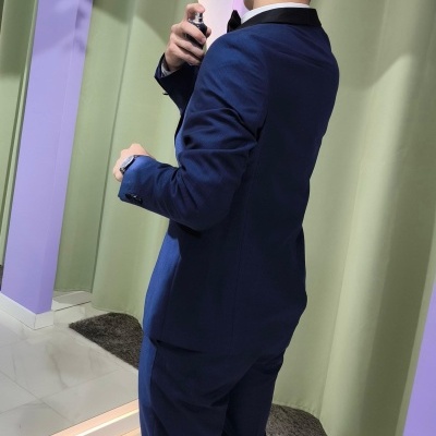 Homem vestido com fato azul a aplicar perfume em cenário moderno com paredes lilás e cortinas verdes