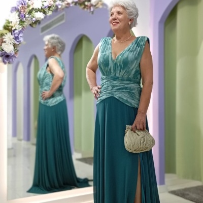 Senhora com vestido longo azul esverdeado em sala com espelho e flores.