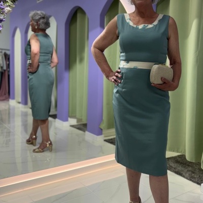 Mulher num vestido azul esverdeado com detalhes bege, sapatos dourados e mala bege, em loja com parede roxa e espelho.