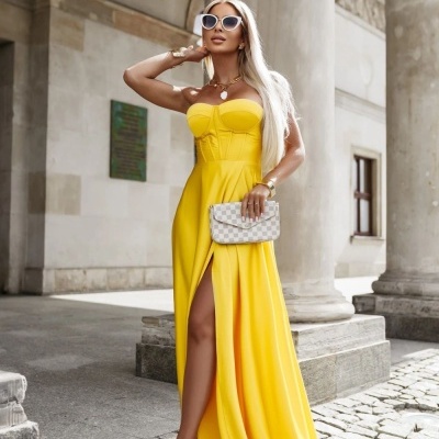Vestido longo amarelo com racha e clutch xadrez em ambiente urbano com pilares de pedra.