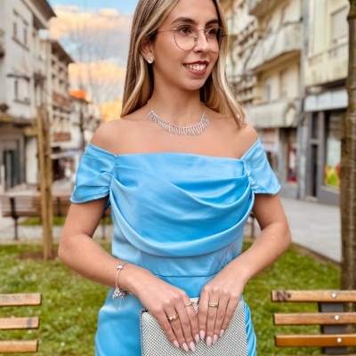 Mulher com vestido azul claro acetinado e clutch prateada numa rua urbana