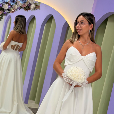 Mulher com vestido de noiva branco, óculos e bouquet em frente a espelho com decoração floral