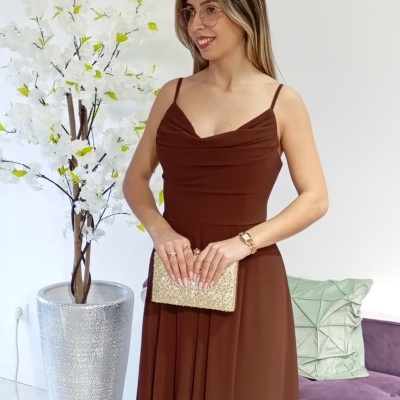 Mulher com vestido castanho usando clutch dourada e acessórios dourados, com planta branca e sofá roxo ao fundo