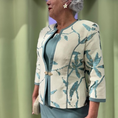 Mulher com casaco estampado de folhas azuis e vestido azul-petróleo em fundo de cortinas roxas e verdes