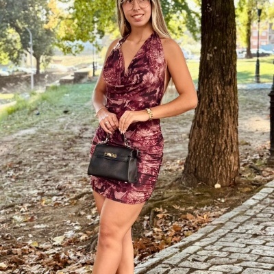 Mulher com vestido roxo estampado, sapatos pretos e mala preta de mão em passeio ao ar livre
