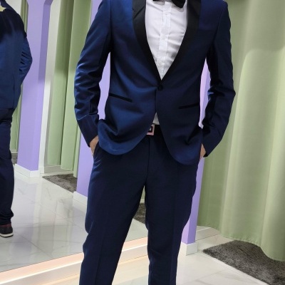 Homem num fato azul escuro com gravata borboleta preta num espaço com arco roxo e flores artificiais.