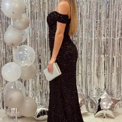 Mulher com vestido preto longo com lantejoulas e clutch prata, fundo prateado com balões.