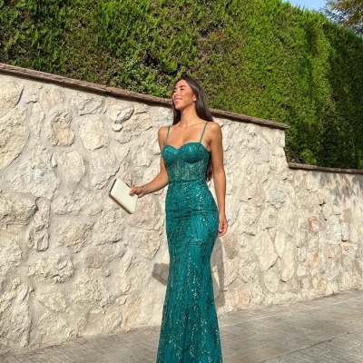 Mulher a usar vestido longo verde com renda e alças finas, segurando carteira bege, junto a parede de pedra e sebes.