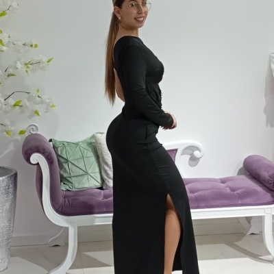 Mulher com vestido preto longo e óculos, ao lado de sofá branco com almofadas coloridas.