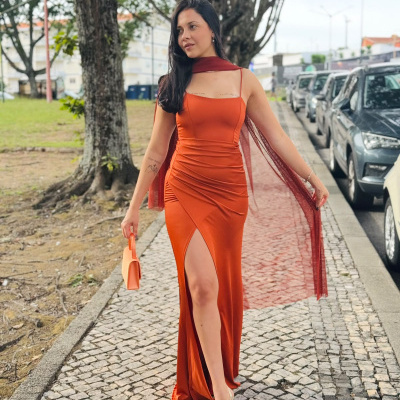 Mulher com vestido laranja longo com fenda e alças finas, segurando mala laranja e andando numa calçada de pedra.