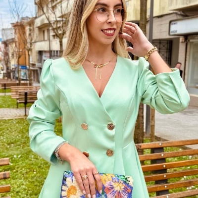 Mulher vestindo vestido verde claro com botões dourados segurando mala de mão floral colorida em parque urbano