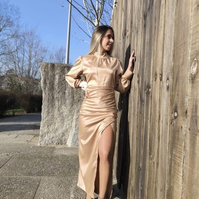 Mulher de vestido comprido dourado com fenda encostada a vedação de madeira