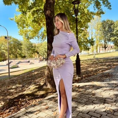 Mulher com vestido lilás longo e clutch bege floral em área exterior com árvores e calçada de pedra