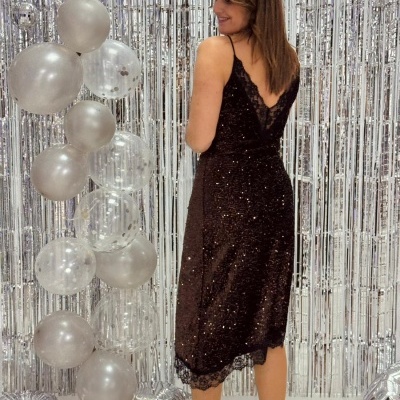 Vestido preto brilhante com alças finas e renda, sapatos rosas, e decoração prateada com balões.