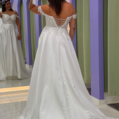 Vestido de noiva branco com corpete em renda e saia longa, visto de costas, mulher segura bouquet branco em sala com parede lilás e verde e espelho.}