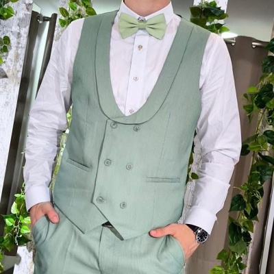 Homem jovem com fato verde-menta, colete, calças, camisa branca e gravata borboleta verde em ambiente com plantas e madeira branca.