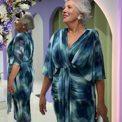 Mulher com vestido azul e verde de manga média, cinto e sandálias nude, junto a espelho decorado com flores lilás