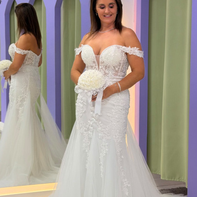 Mulher com vestido de noiva branco e bouquet, frente a espelho com moldura lilás e cortinas verdes