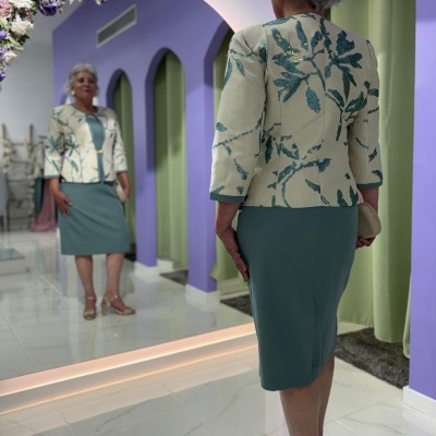 Mulher idosa veste vestido verde e casaco bege com padrão vegetal junto a espelho em espaço lilás com arco de flores