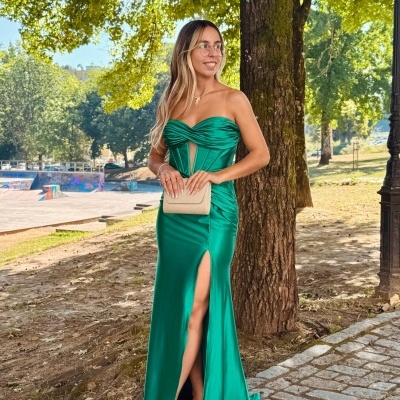 Mulher em vestido longo verde esmeralda posando ao ar livre junto a árvore e calçada de pedra