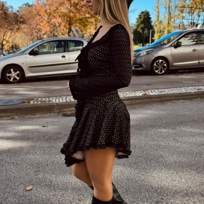 Mulher com vestido preto de pontos brancos e botas pretas numa rua com carros e árvores