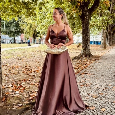 Mulher com vestido longo castanho e mala dourada num parque de árvores em outono
