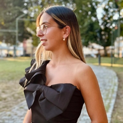 Mulher de perfil com vestido preto com laço largo, óculos dourados e brincos de coração ao ar livre