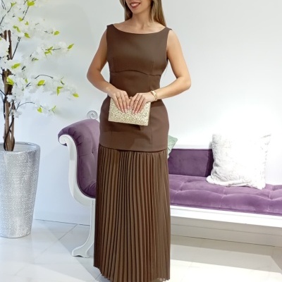 Mulher com vestido castanho comprido plissado e clutch dourada ao lado de sofá roxo e vaso com flores brancas