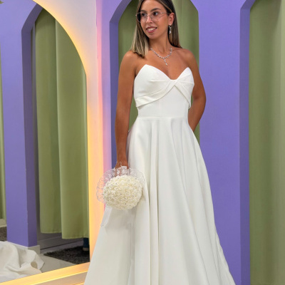 Vestido de noiva branco com corpete tomara que caia e saia ampla, bouquet de flores, mulher com joias e óculos em fundo com arcos violetas.
