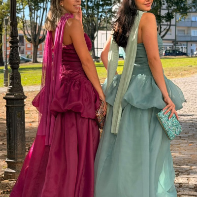 Mulheres com vestidos longos de organza vinho e azul claro com faixas no pescoço segurando carteiras brilhantes em calçada de pedras