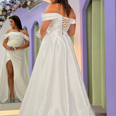 Vestido de noiva branco com ombros descobertos e amarração nas costas, refletido num espelho com decoração floral e parede lilás com arcos verdes.