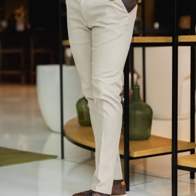 Calças bege com cinto castanho, camisa branca e sapatos castanhos em ambiente interior com móvel decorativo