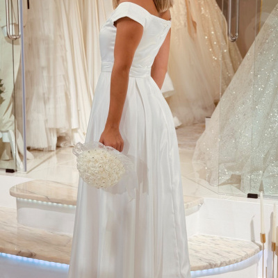 Vestido de noiva branco de tecido acetinado com corte ombro a ombro e bouquet branco em loja