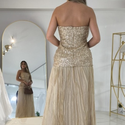 Vestido longo dourado com corpete brilhante e saia plissada bege em ambiente interior