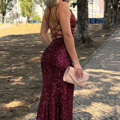 Mulher de vestido longo bordô brilhante segurando clutch rosa numa calçada de pedra ao ar livre.