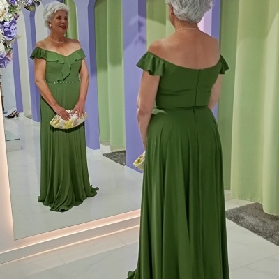 Senhora com vestido longo verde e carteira dourada em frente a espelho decorado com flores.