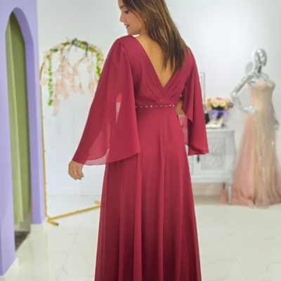 Vestido longo vinho com mangas largas e decote V nas costas numa sala clara com chão de mármore.