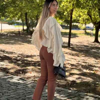 Mulher com camisola branca, calças marrons, ténis brancos com padrão castanho, e mala preta ao ar livre.