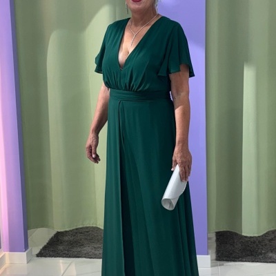 Mulher com vestido verde escuro longo e carteira branca em interior com paredes lilás.