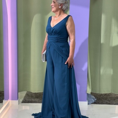 Mulher com vestido comprido azul petróleo e carteira prateada em fundo de cortinas verdes e paredes lilás.