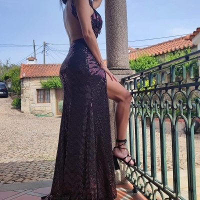 Mulher com vestido longo preto brilhante e fenda, encostada numa coluna de pedra numa varanda