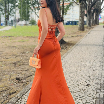 Modelo usa vestido longo laranja de cetim com alças finas e mala de mão laranja ao ar livre.