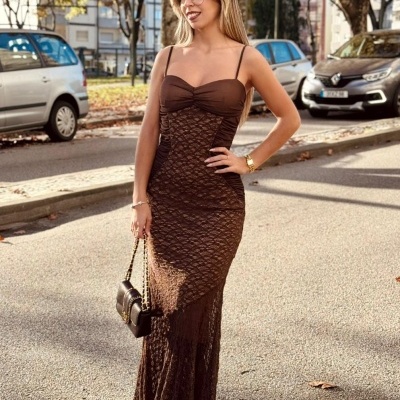 Mulher com vestido comprido castanho e mala de mão castanha num ambiente urbano de rua com árvores e carros.