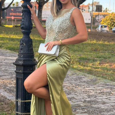 Mulher com vestido longo verde e pedras, segurando mala prateada ao lado de poste preto