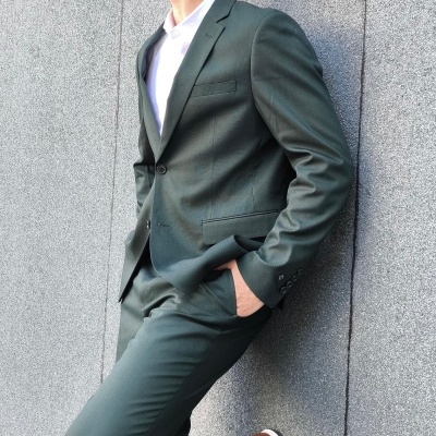 Homem com fato verde escuro, camisa branca e sapatos castanhos encostado numa parede cinzenta