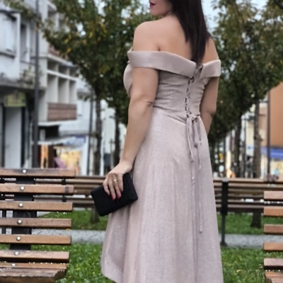 Mulher de costas usando vestido rosa claro ombros fora, sapatos pretos e carteira preta num parque urbano.