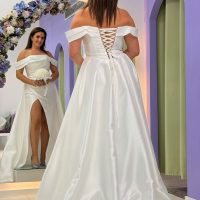 Vestido de noiva branco em tecido acetinado com decote ombro a ombro e costas entrelaçadas, com saia longa e racha frontal